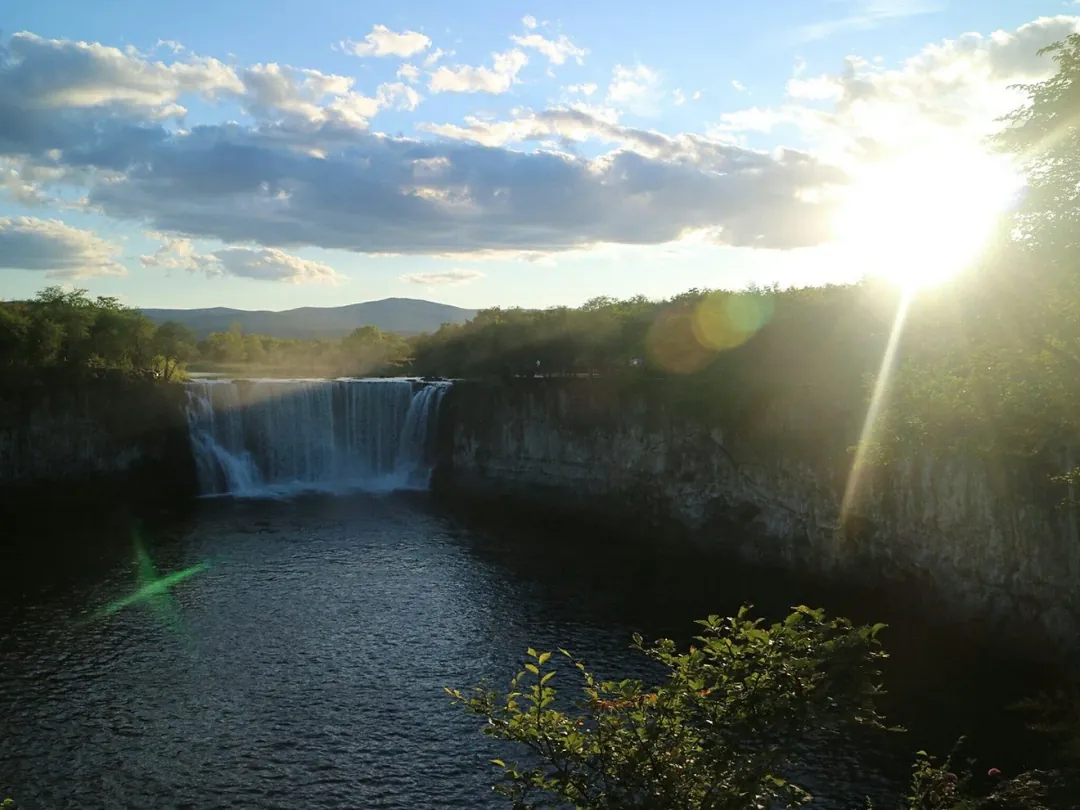 Diaoshuilou Waterfall at sunset: the Mudanjiang river pouring over a dark basalt lip into the pool below, sun flaring through the trees