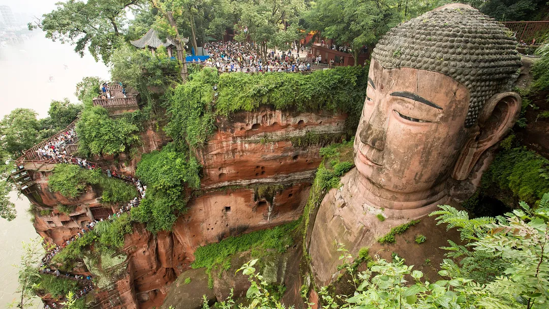 Close-up of the Buddha's head with the nine-turn plank road and a queue of visitors on the cliff beside it