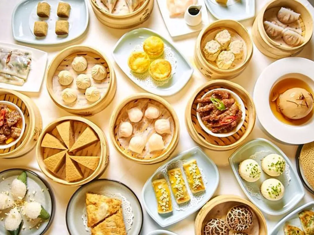 An overhead spread of dim sum: bamboo steamers of har gow and shumai, rice rolls and egg tarts on white plates