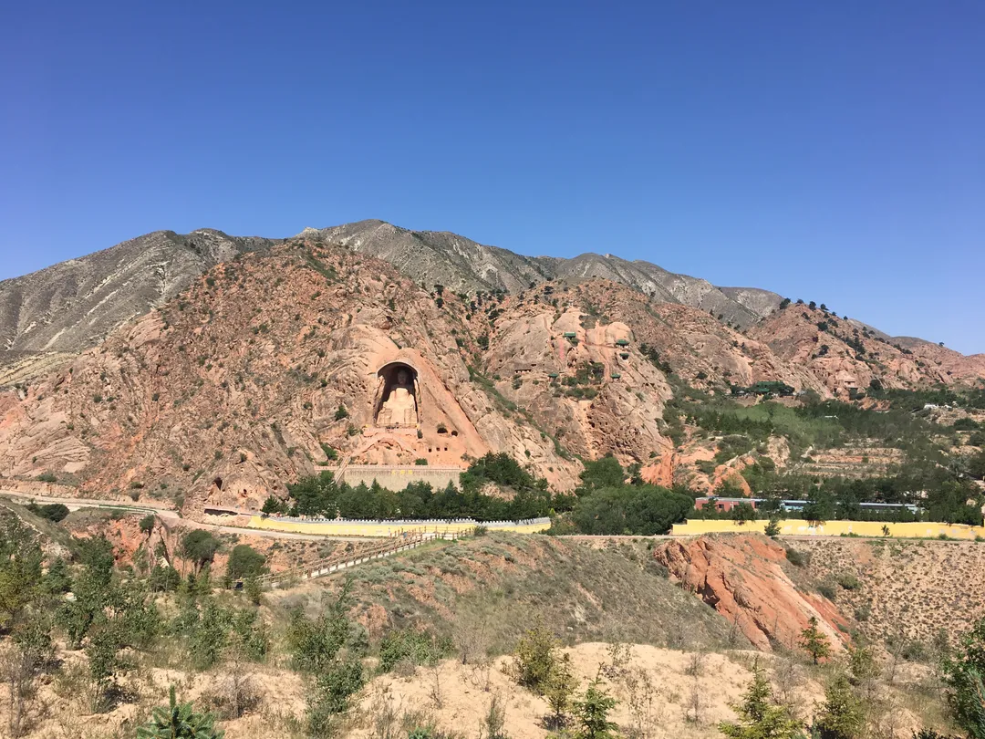 The Xumishan cliff with the giant seated Buddha niche carved into the red sandstone, seen across a dry valley with the site road below