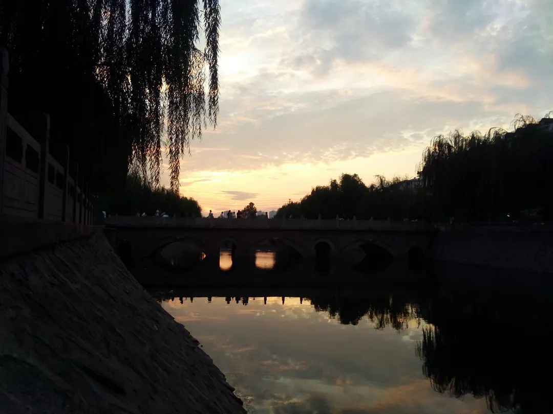 El puente de piedra de Xuebu con sus arcos recortado al atardecer sobre el viejo cauce, sauces en ambas orillas y reflejos en el agua