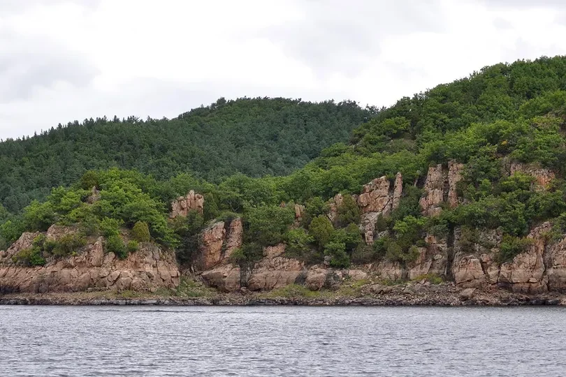 Rocky bluffs and green forest running into the calm water of Jingpo Lake under a grey sky